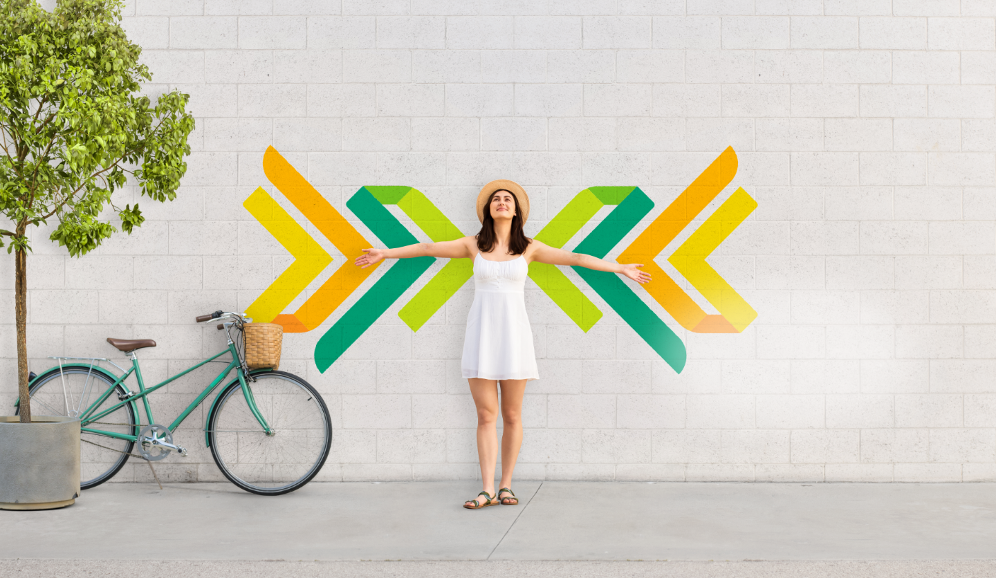 A woman in a white dress standing with open arms before a colorful chevron mural, beside a bicycle and tree